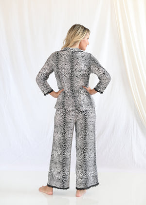 A woman stands with her back to the camera, hands on her hips, wearing a Shapeez Women's Leopard Print Bamboo Lounge Pajamas Set with 3/4 Sleeve Top and Pants against a white curtain backdrop.